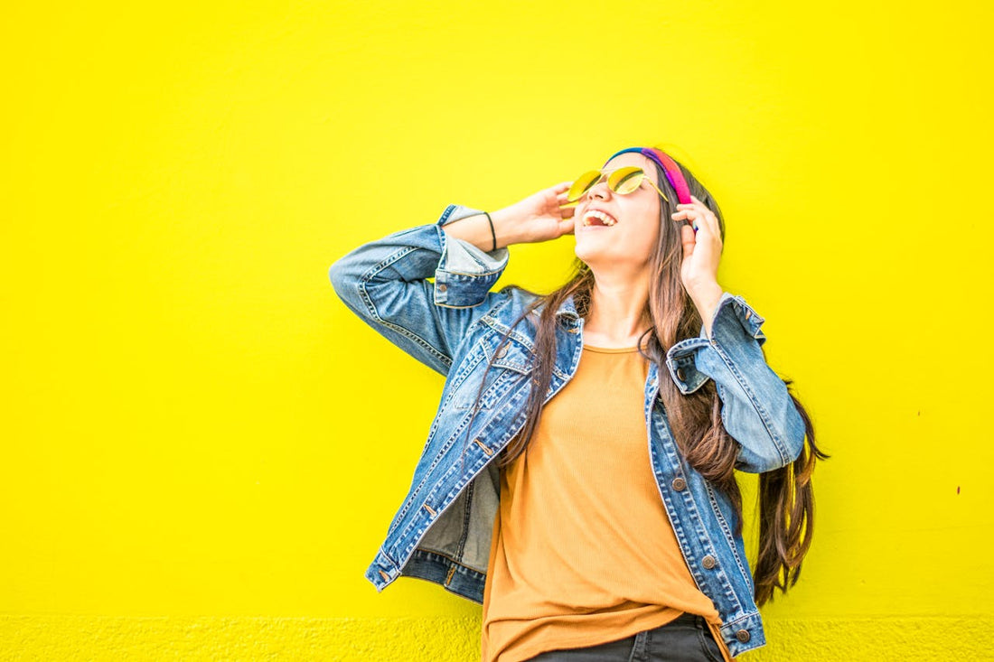 A cheerful woman wearing a denim jacket, mustard yellow top, and yellow sunglasses smiles while standing against a bright yellow wall. She is also wearing a colorful headband and has her hands raised to adjust her sunglasses