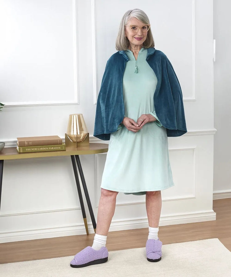Senior woman wearing a luxurious teal sleep cape over a light mint green dress, complemented by cozy lavender grip slippers, posing in a stylish home setting