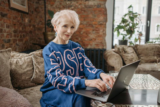 Caucasian women with white hair, a blue sweater with pink details typing on a computer.