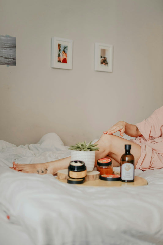 Person in a pink robe applying skincare to their legs while relaxing on a white bed, with an assortment of body care products and a small succulent plant arranged on a wooden tray nearby.