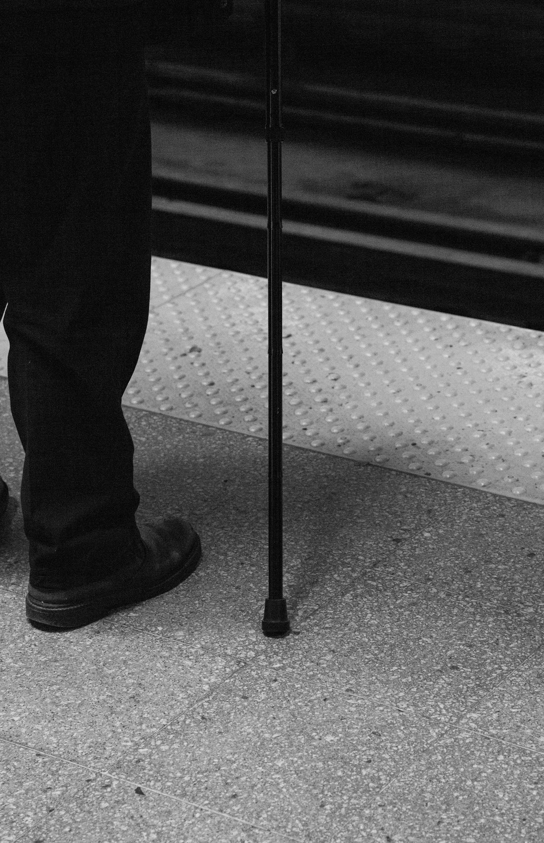 Black and white photo of a person standing on a subway platform, wearing dark pants and shoes, and using a mobility cane
