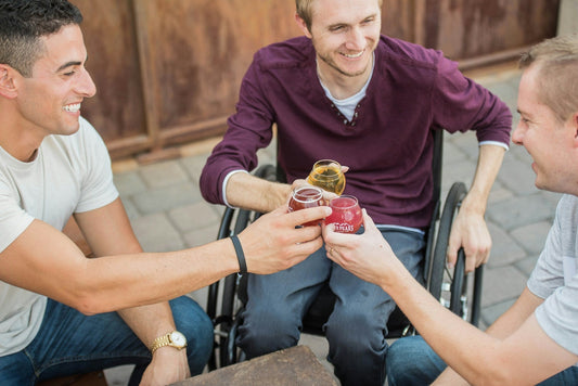 Three men smiling and toasting with drinks while sitting outside. One man is in a wheelchair, and all appear to be enjoying a casual, friendly gathering.