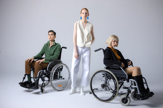 Three fashion models pose in a studio setting—two seated in wheelchairs and one standing between them—wearing stylish, modern adaptive clothing