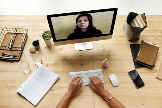 A person types on a white keyboard during a video call with a woman shown on a desktop monitor. The wooden desk is organized with notebooks, pens, a water bottle, a smartphone, a mouse, small potted plants, and a wire organizer.