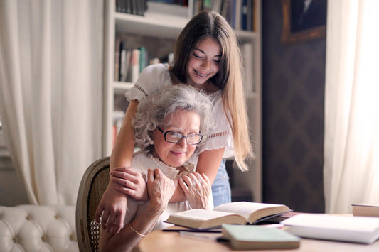 A smiling elderly woman wearing glasses reads a book at a table while a young girl lovingly hugs her from behind. They are in a cozy, well-lit room with bookshelves