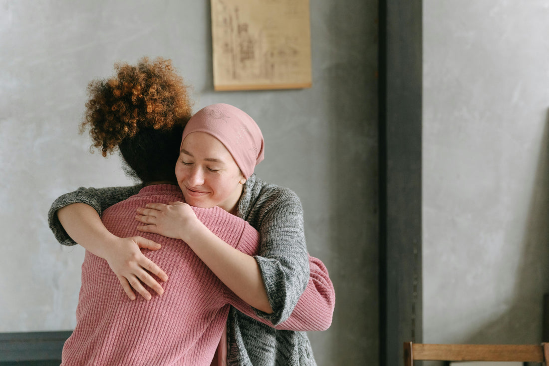 One women hugging another for comfort, one women is wearing a pink blouse with curly auburn hair where the other is wearing a green top with a pink cover over her head