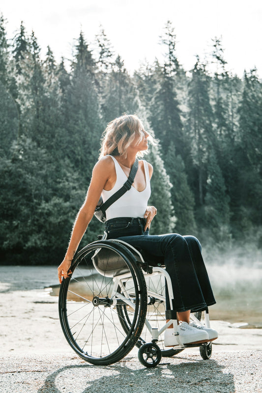 A woman with short blonde hair sits in a white manual wheelchair outdoors, wearing a white tank top, dark jeans, and white sneakers. She is smiling and looking to the side, with a forest and misty lake in the background on a sunny day