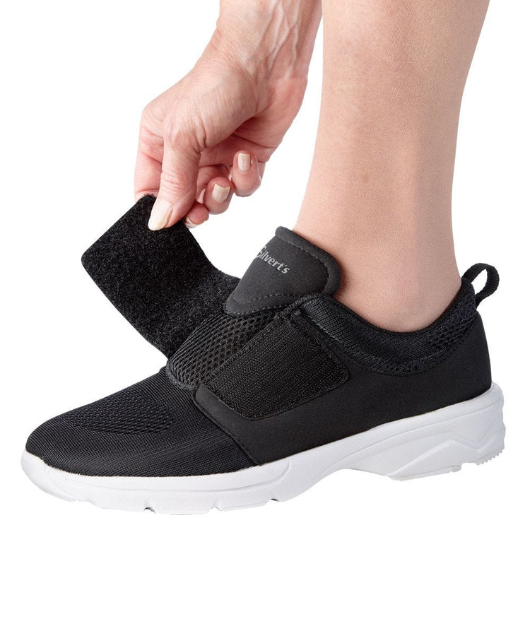 Women putting on black wide walking shoes with white bottom and velcro closure.